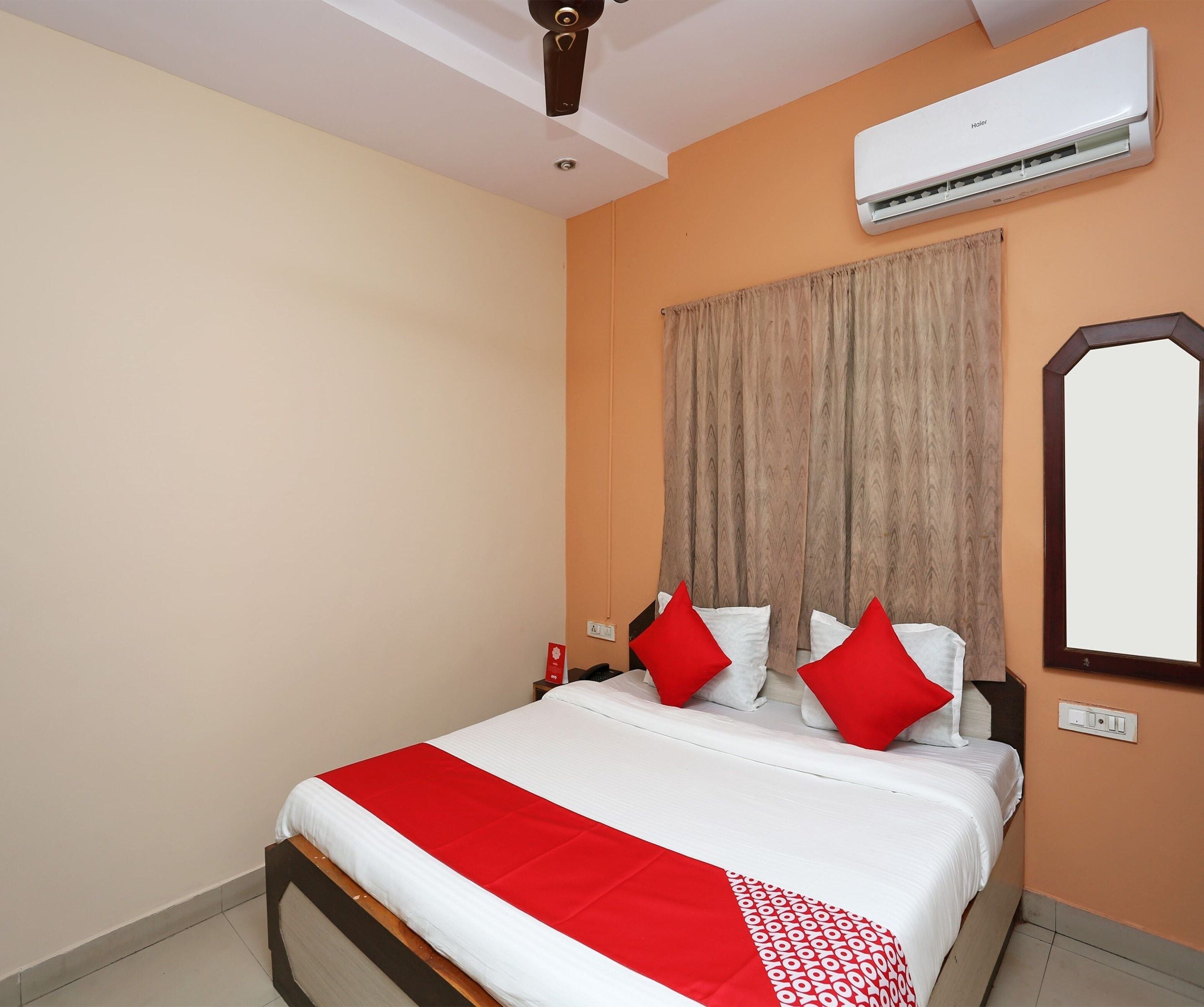 A hotel room with a bed, a ceiling fan, an air conditioning unit, a wall-mounted mirror, and a window with curtains.