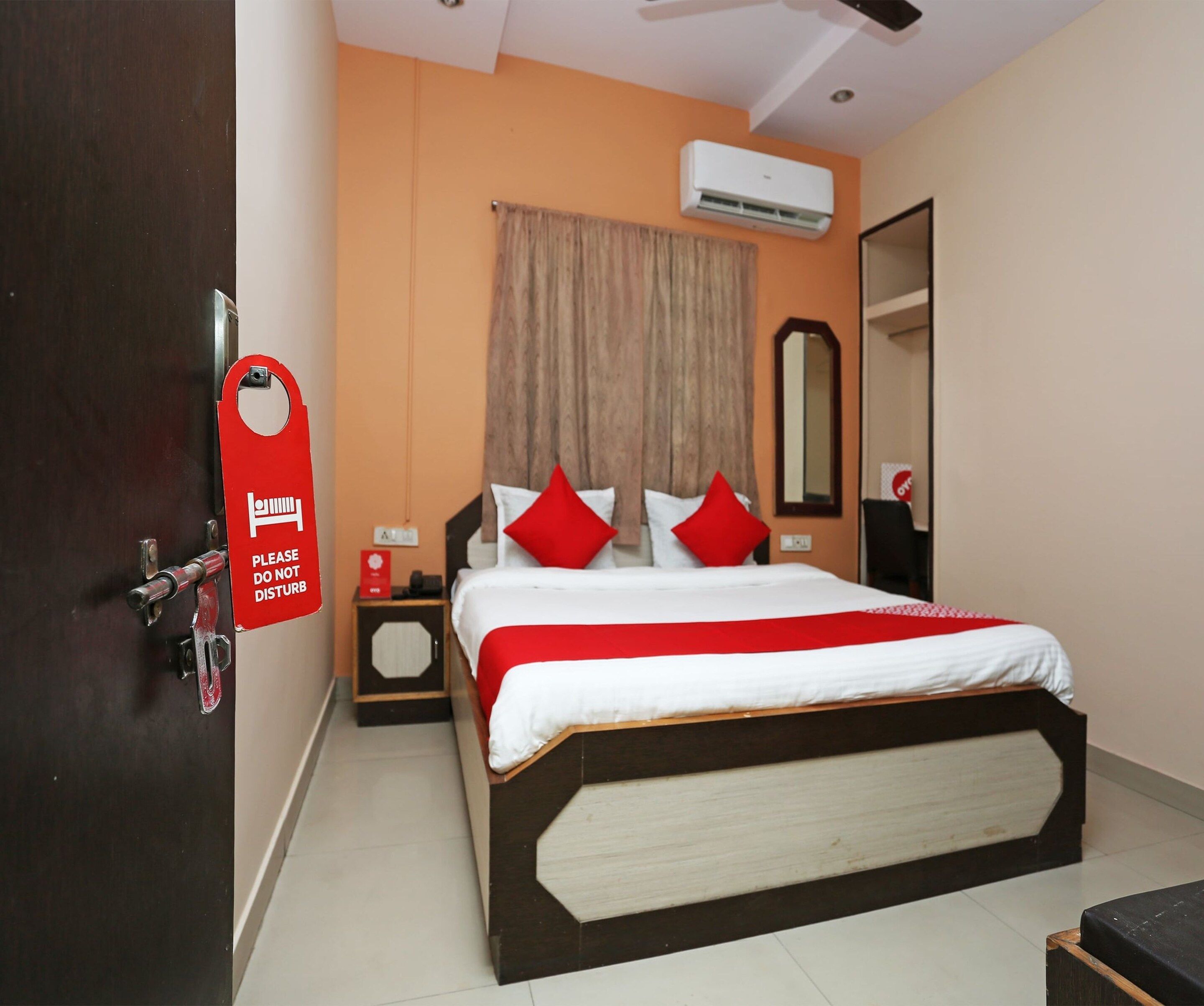 A hotel room with a bed, a nightstand, an air conditioning unit, a mirror, and a door with a 'Please Do Not Disturb' sign.