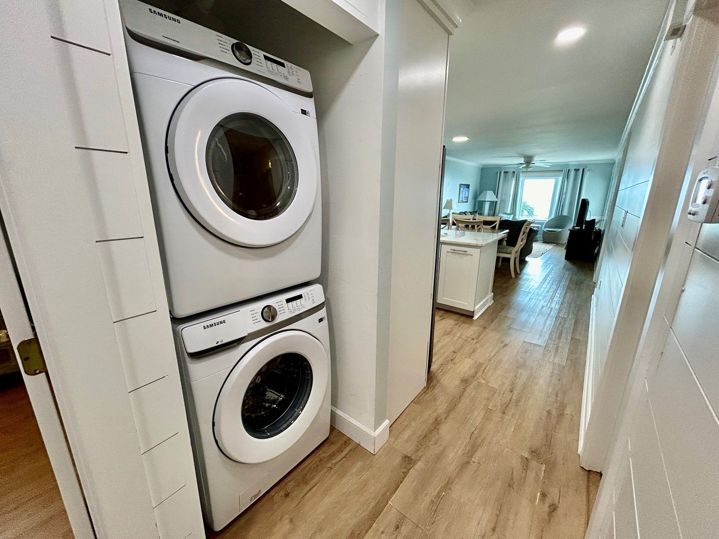 A laundry area with two stacked washing machines, a wooden floor, and a view into a living room area with a dining table and chairs.