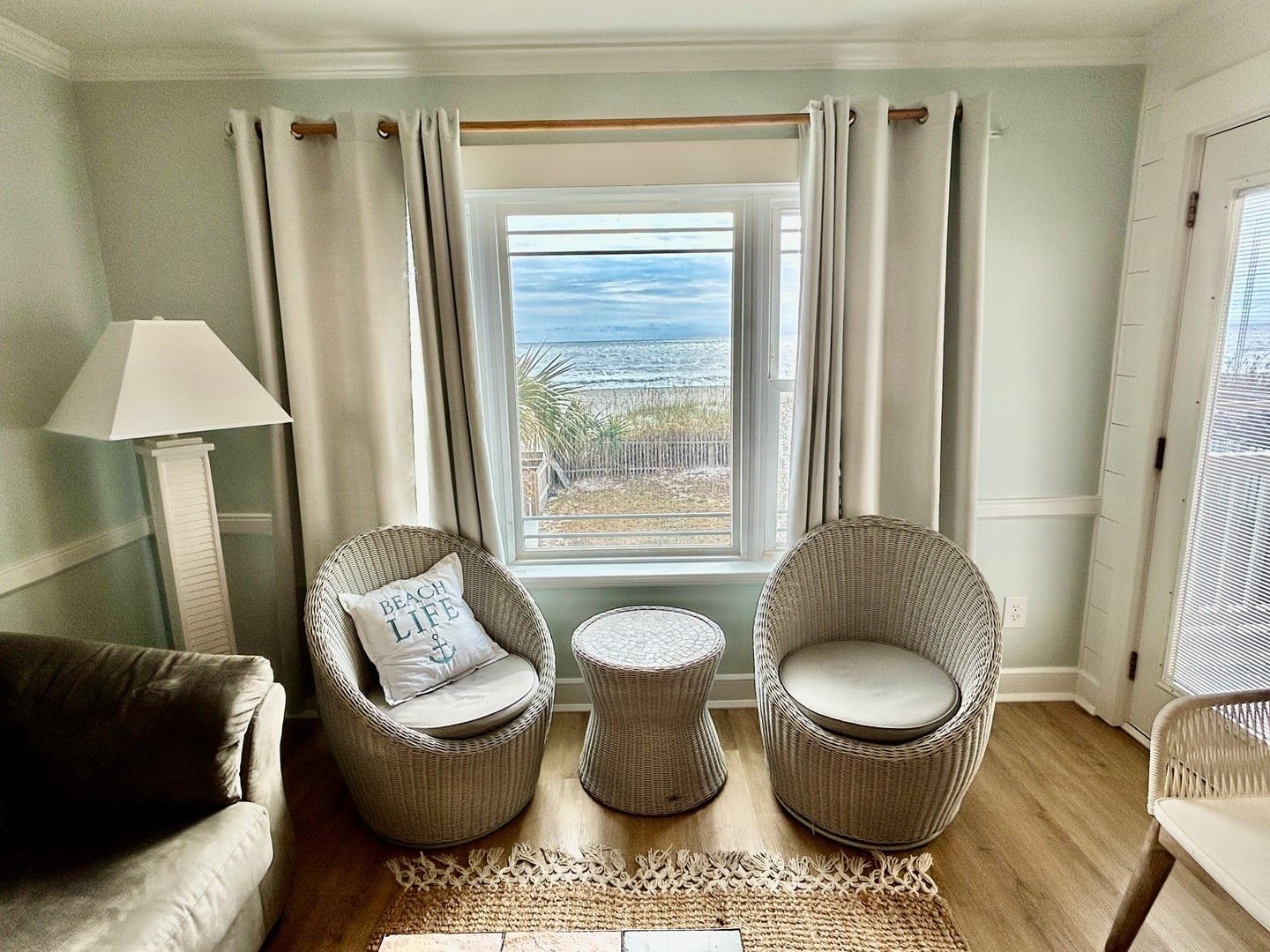 A room with a view of the ocean, two wicker chairs with cushions, a small round table, and a floor lamp.