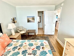 Ein Schlafzimmer mit einem Bett, einem Sessel, einem Nachttisch, einer Lampe und einer Tür, die in einen Flur führt.