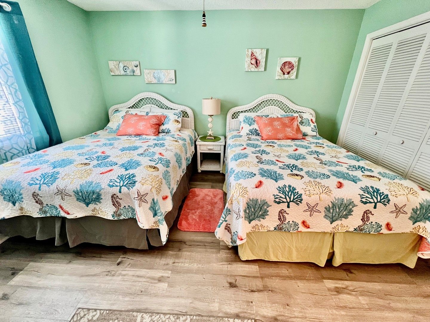 Una habitación con dos camas, cada una con una colcha estampada, una mesita de noche con lámpara y una ventana con contraventanas.