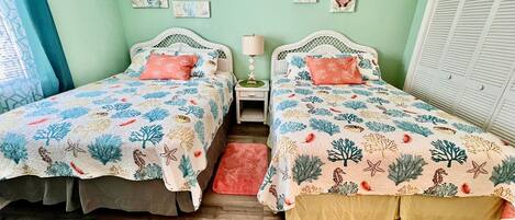 Una habitación con dos camas, cada una con una colcha estampada, una mesita de noche con lámpara y una ventana con contraventanas.