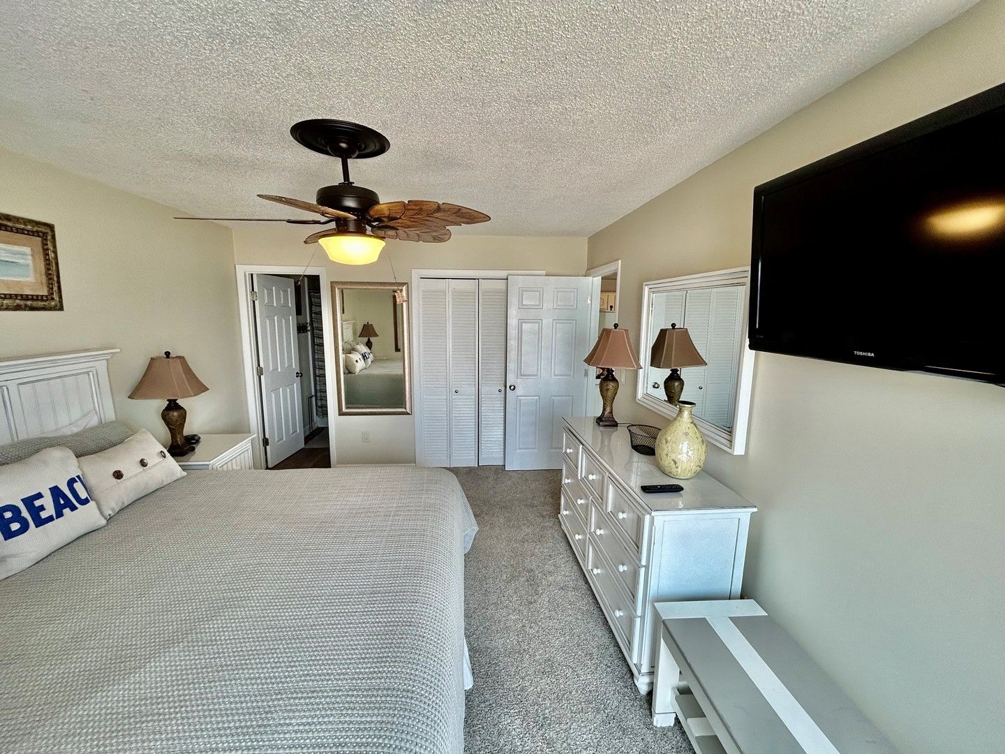 A bedroom with a bed, a dresser, a mirror, a ceiling fan, and a TV mounted on the wall.