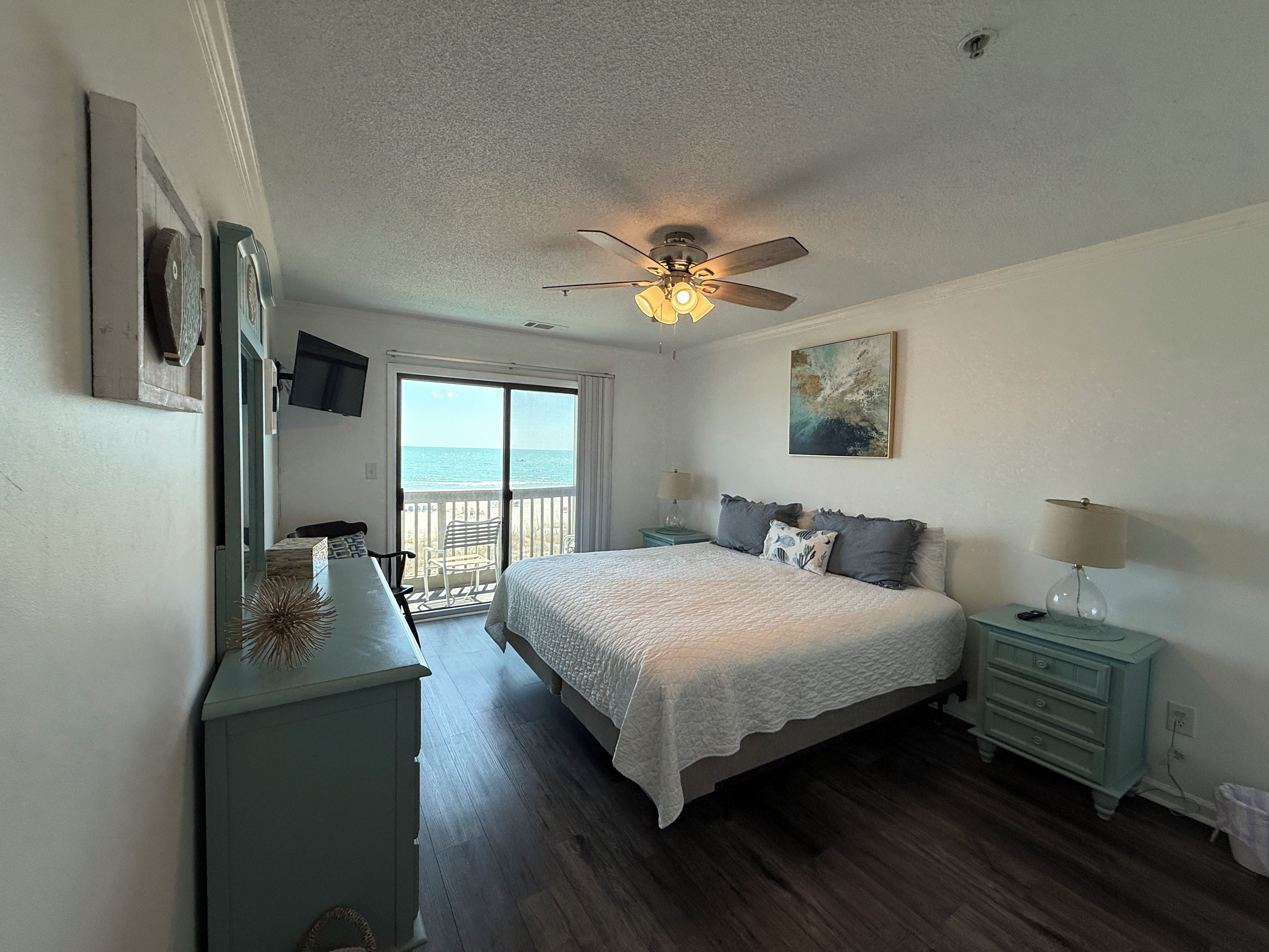 A bedroom with a bed, a ceiling fan, a nightstand, a dresser, and a view of the ocean through a sliding door.