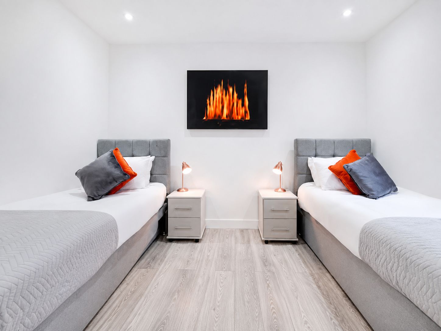 A modern hotel room with two beds, a nightstand with a lamp, a wall-mounted artwork of flames, and a light-colored wooden floor.