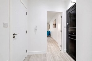 A hallway with white walls, a wooden floor, and a black door on the right. There is a blue chair in the distance and a phone on the wall. - Contractor/CorporateTravel/Relocation, MyRent2Rent (Corby)