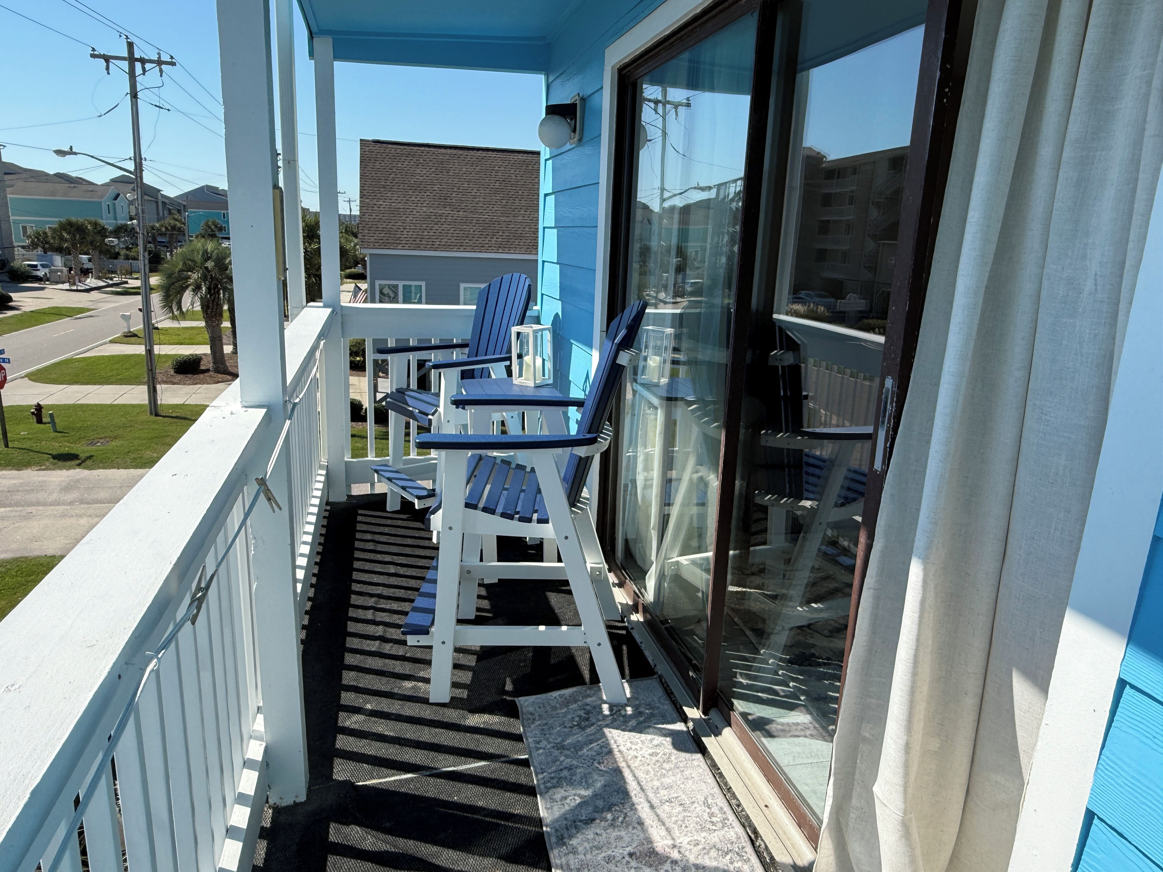 A balcony with two blue chairs and a table, overlooking a street with houses and a clear sky.