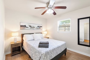 A bedroom with a bed, a ceiling fan, a nightstand, a lamp, a window, and a wall-mounted mirror. - Tampa 3BR Near Airport & MacDill | Sleeps 6 (Tampa)
