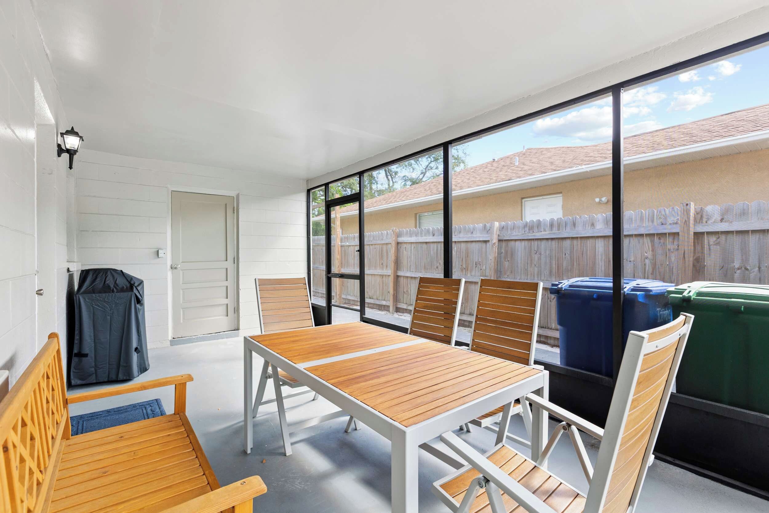 Eine Terrassenfläche mit Tisch und Stühlen, eine überdachte Sitzbank und Blick auf einen Garten mit Holzzaun und Mülltonnen.