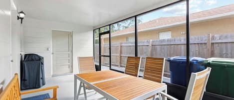 A patio area with a table and chairs, a covered bench, and a view of a backyard with a wooden fence and trash bins.