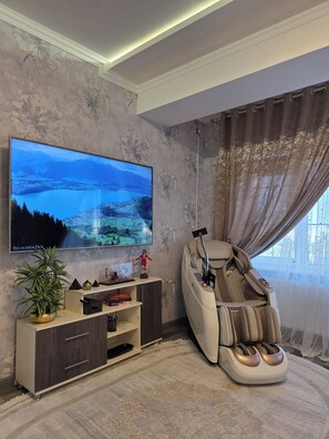 A living room with a flat-screen TV mounted on the wall, a massage chair, a potted plant, and a small shelf with decorative items.