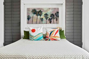 Ein Bett mit gemusterten Betttextilien, zwei Kissen mit verschiedenen Designs und ein an der Wand hängendes Bild von Palmen.