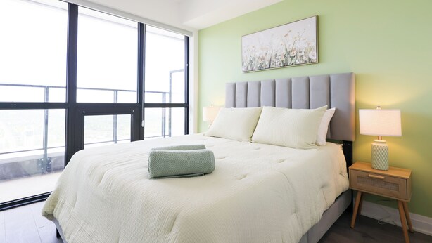A bedroom with a large bed, a nightstand with a lamp, and a window with a view. - Casa Verde | Skyline Views Near Square One (Mississauga)