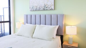 A bed with a grey upholstered headboard, two white pillows, and a white textured bedspread. There is a bedside table with a lamp and a water bottle on it. A framed floral artwork is above the bed. - Casa Verde | Skyline Views Near Square One (Mississauga)