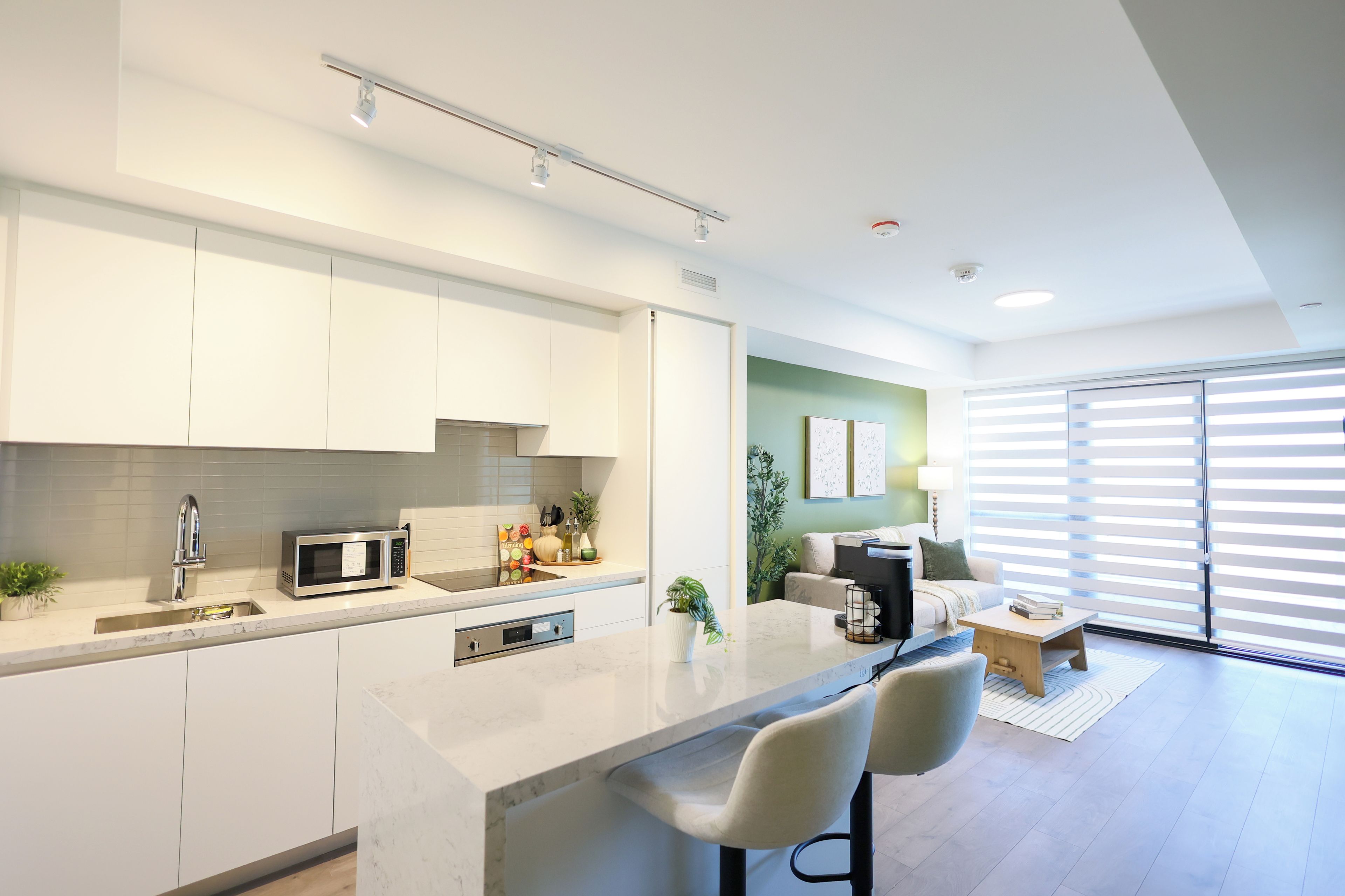 A modern kitchen with a built-in microwave, a sink, and a countertop. There is a dining area with a sofa and a coffee table. The room has a large window with blinds.