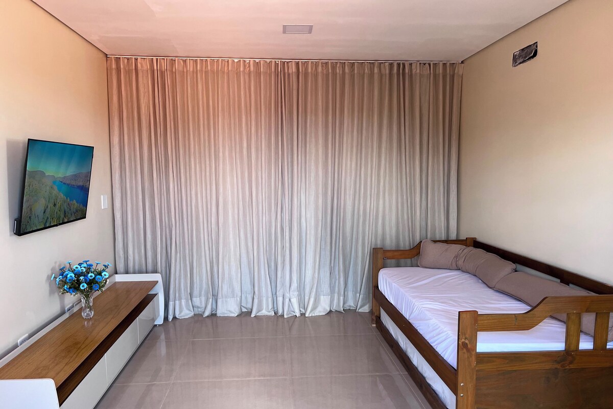A bedroom with a bed, a bedside table with a vase of flowers, a wall-mounted TV, and a large window with curtains.