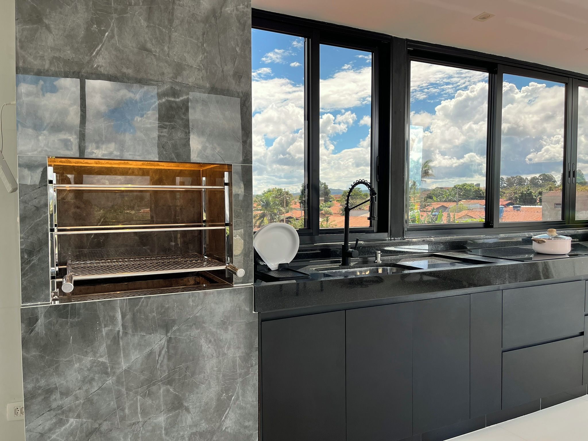 Une cuisine moderne dotée d’un barbecue intégré, d’une grande fenêtre offrant une vue sur le ciel et les nuages, et d’un évier avec un robinet.