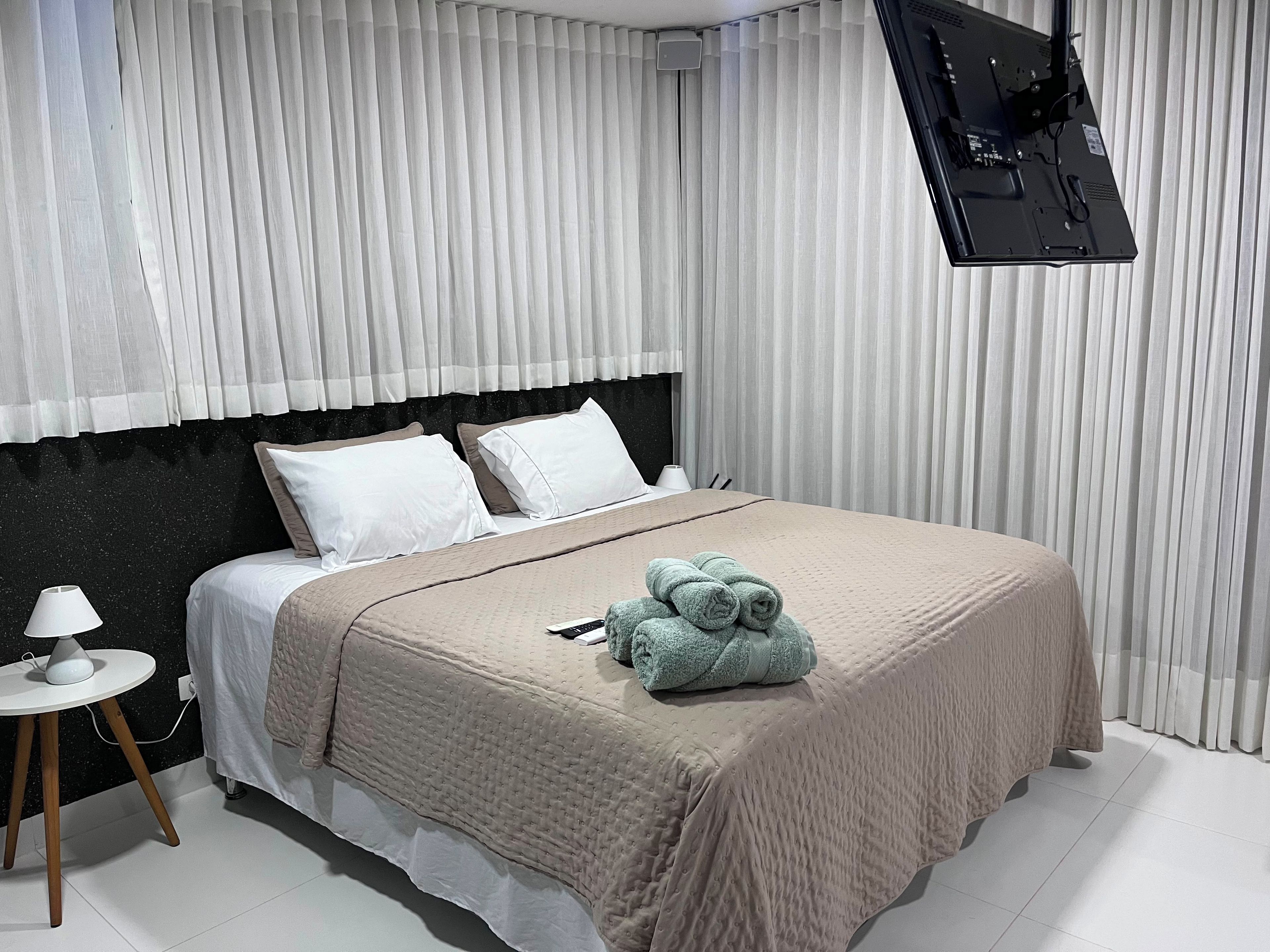 Un lit bien fait, recouvert d’une couverture beige, avec des oreillers et une serviette enroulée posée sur le lit. Une petite lampe blanche se trouve sur une table de chevet à côté du lit.