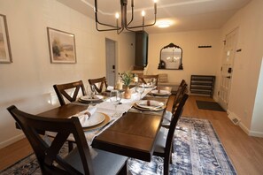 Une salle à manger avec une longue table dressée pour six personnes, des chaises en bois, un lustre et un miroir au-dessus de la table.