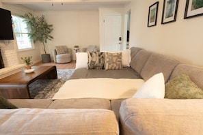 A modern living room with a large sofa, a coffee table, and a television. There is a potted plant and a door in the background. - Modern Home - Lux 5 Bdrm/3.5 Bath Sleeps 14 (Grandview)