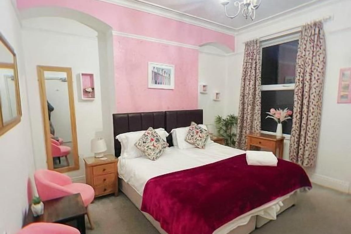 A bedroom with a bed, a chair, a nightstand, a mirror, and a window with curtains.