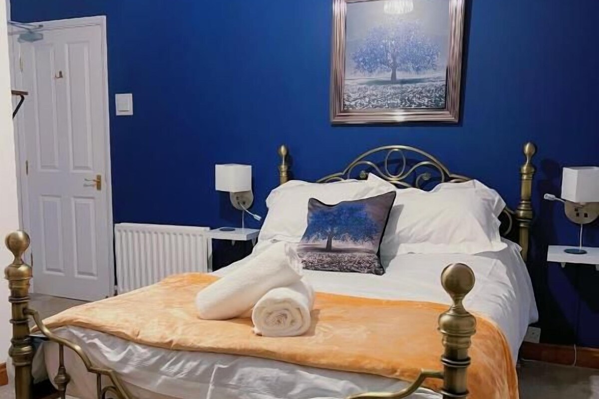 A bedroom with a bed, a white door, a framed picture on the wall, and a bedside table with a lamp.