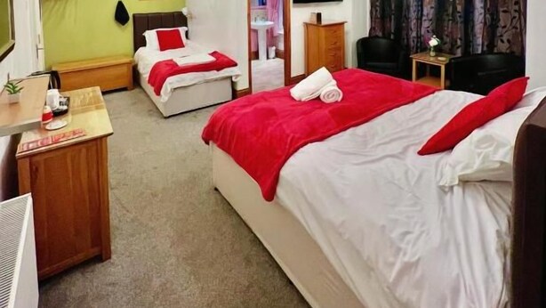 A hotel room with two beds, a desk, a chair, a TV, and a window with curtains. - Ellie's Guest House (Whitby)