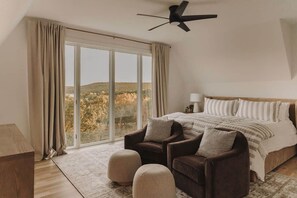 A bedroom with a large bed, two armchairs, a ceiling fan, and a view of a hilly landscape through a large window. - Lakefront Retreat | The Lucas (Eufaula)