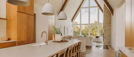 A kitchen with a large island, wooden cabinets, pendant lighting, and a view through a large window.