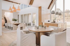 A spacious living area with a round table and white chairs, a kitchen with wooden cabinets, and large windows offering a view of the outdoors. - Lakefront Retreat | The Lucas (Eufaula)