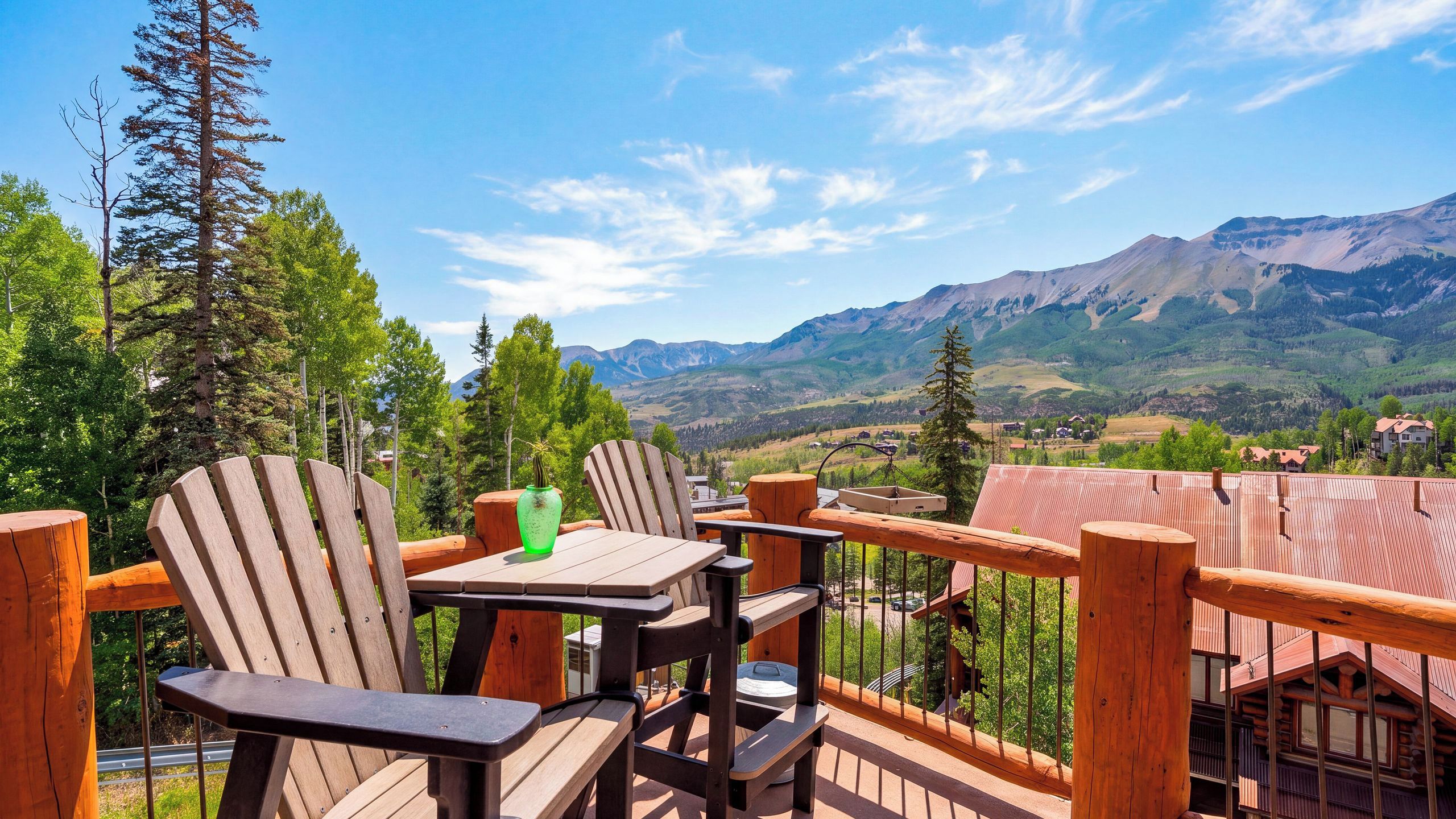 A wooden deck with Adirondack chairs and a table, overlooking a mountainous landscape with houses in the valley.