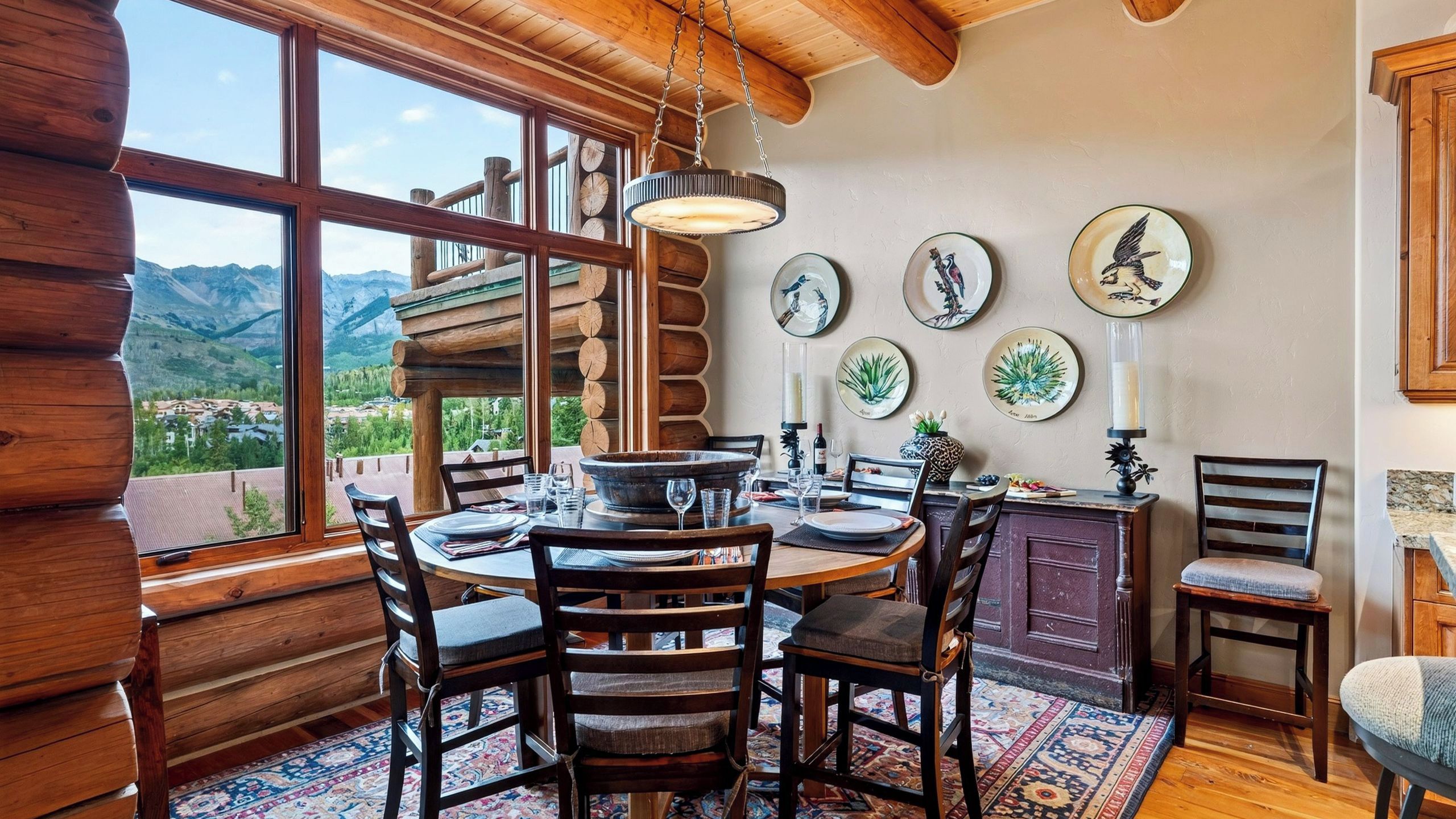 A dining area with a wooden table and chairs, a view of mountains, and decorative plates on the wall.