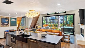 A modern kitchen with a central island, a dining area with a sofa and table, and a large window with a view of trees and a building. - The Blackwood A-frame by Avantstay Hot Tub & Pond Arcade Room EV Charger Near Slopes & Lake (Big Bear Lake)