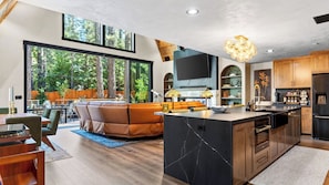 A modern kitchen with a marble island, wooden cabinets, and a large window overlooking a forest. - The Blackwood A-frame by Avantstay Hot Tub & Pond Arcade Room EV Charger Near Slopes & Lake (Big Bear Lake)