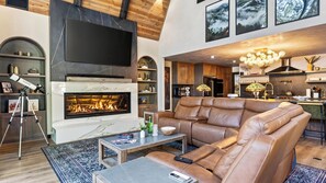 A modern living room with a fireplace, leather sofa, and a dining area with wooden cabinets. - The Blackwood A-frame by Avantstay Hot Tub & Pond Arcade Room EV Charger Near Slopes & Lake (Big Bear Lake)