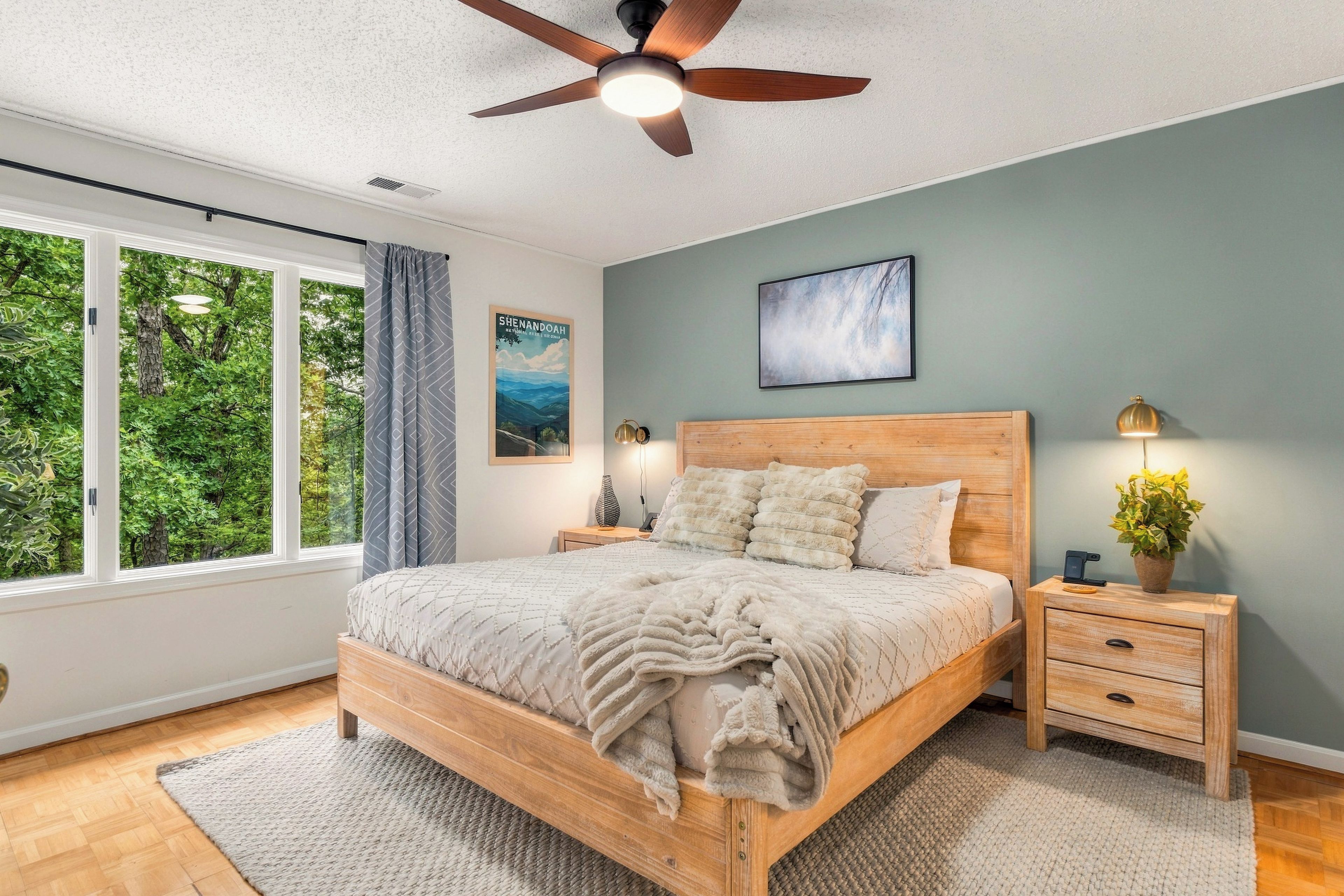 A bedroom with a wooden bed, a nightstand with a lamp, a ceiling fan, a window with curtains, and a wall-mounted picture.