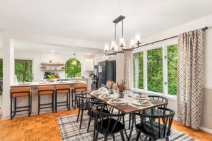 Une cuisine moderne dotée d’un îlot central, d’un coin repas avec une table et des chaises en bois, et de grandes fenêtres offrant une vue sur la verdure.