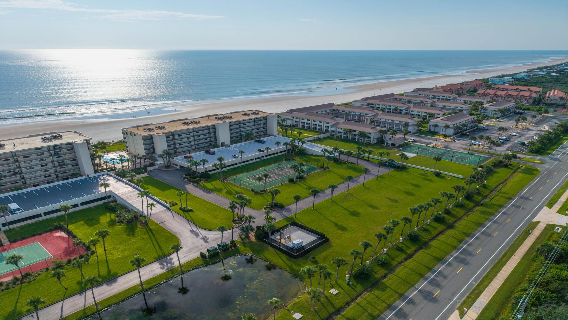 Vue aérienne d’un hôtel situé sur la côte, composé de plusieurs bâtiments, avec une plage et un court de tennis.