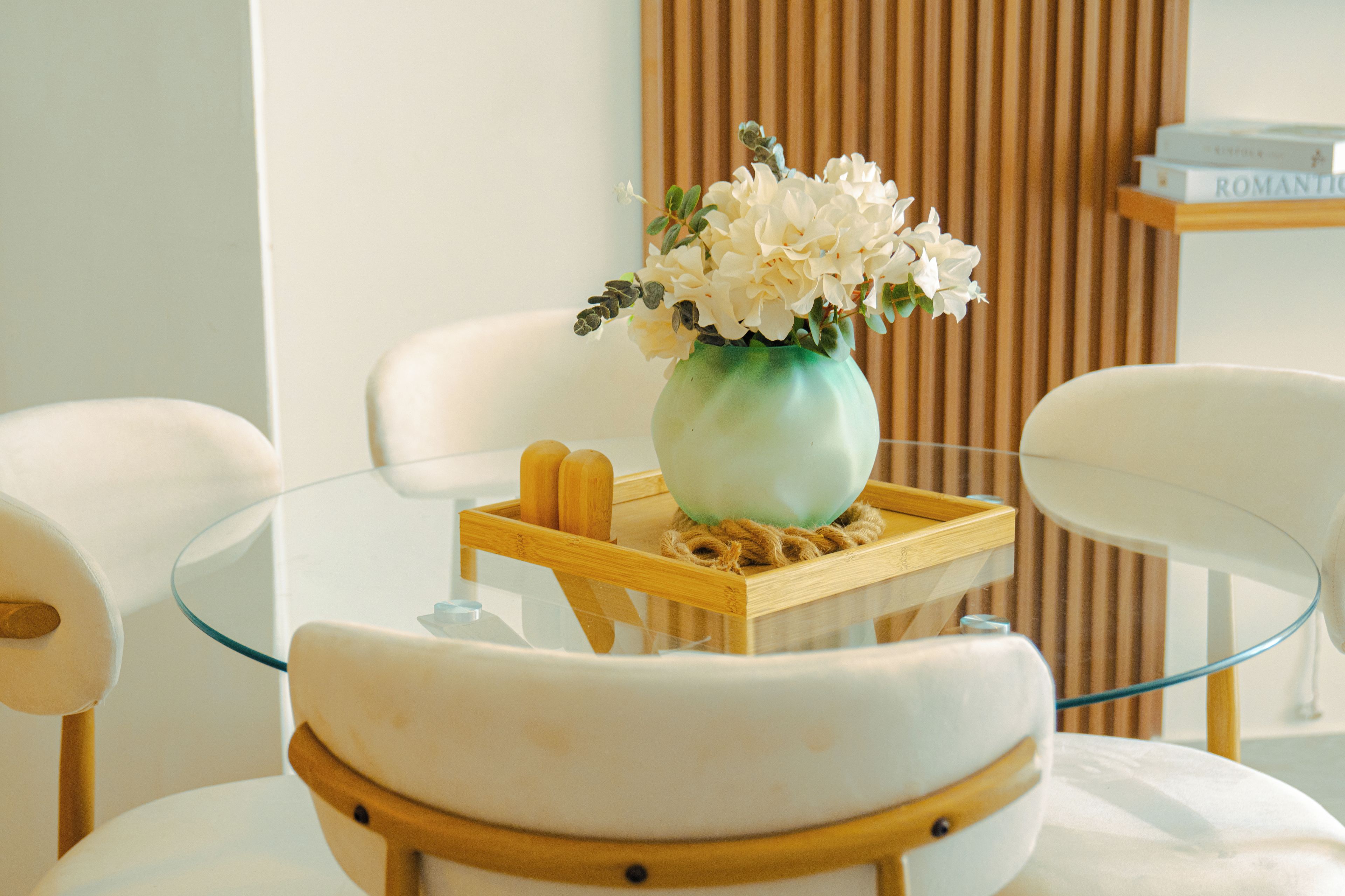 A modern room with a glass table, white chairs, a wooden tray with a vase of flowers, and a bookshelf in the background.