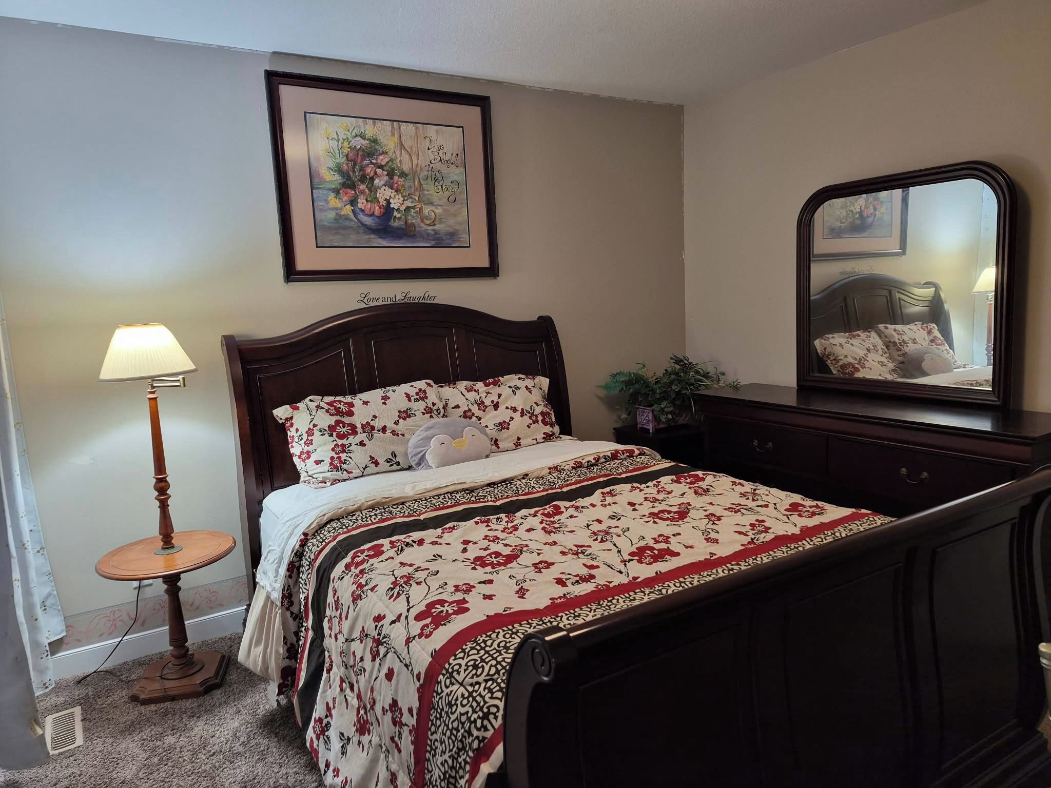 Un dormitorio con cama, lámpara de noche, cómoda, espejo y un cuadro en la pared.