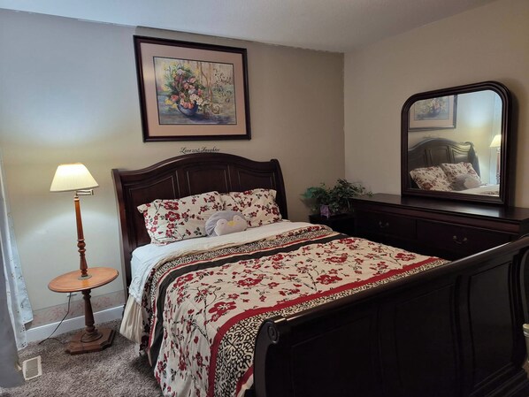 A bedroom with a bed, a bedside lamp, a dresser, a mirror, and a framed picture on the wall. - Home Sweet Home Away From Home (Red Deer)