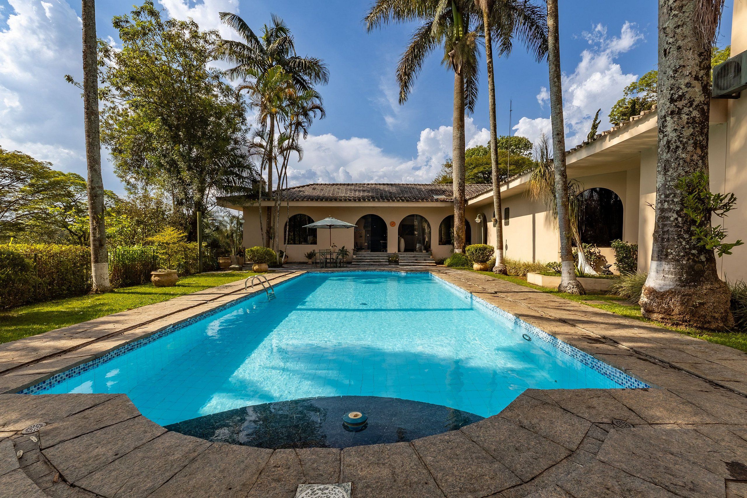 Una piscina con borde de azulejos, rodeada de palmeras y un edificio con ventanas arqueadas.