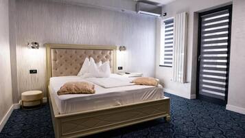 A hotel room with a large bed, a round stool, a wall-mounted light, and a window with blinds.
