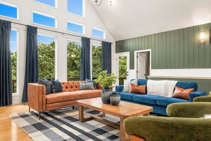 Ein geräumiges Wohnzimmer mit einer Couch, einem Couchtisch und einem blauen Sofa. Große Fenster mit Vorhängen, Blick ins Grüne und eine Tür zu einem anderen Raum.