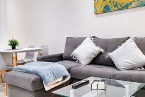En moderne stue med en grå sofa, et blåt plaid, et glas sofabord, en fjernbetjening og en lille plante.