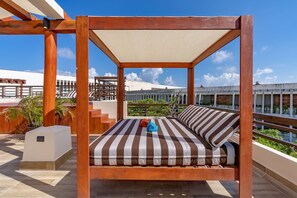 A wooden four-poster bed with striped bedding, a small table, and a chair, set on a balcony with a view of a building and greenery. - Private Pool on Luxury Penthouse (Playa del Carmen)