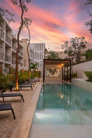 A swimming pool with lounge chairs and a pergola, surrounded by a landscaped garden and a multi-story building. - Private Pool With Great Comfort (Tulum)