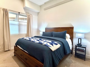 A bedroom with a bed, a nightstand, a lamp, a window with curtains, and a ceiling vent.