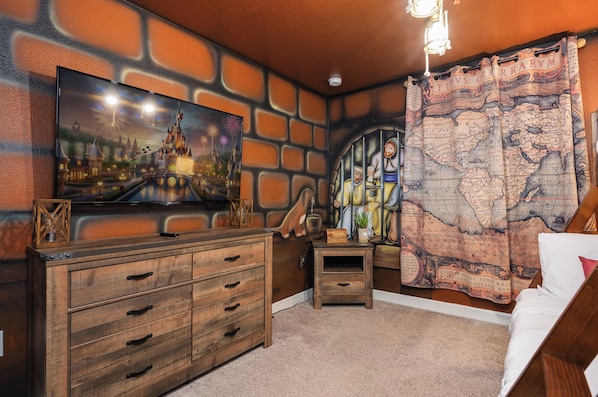 Ein Zimmer mit einem großen Flachbildfernseher an einer Ziegelwand, einer Holzkommode mit mehreren Schubladen, einem kleinen Tisch mit einer Dekoration und einem Vorhang mit Kartenmotiv.